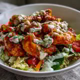 In a white bowl, tender Buffalo Chicken Bowl pieces glisten with sauce beside shredded carrots, cucumber, and cherry tomatoes, ready to be enjoyed. 