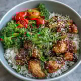 Bite-sized Honey Garlic Chicken glazed in sticky sauce is topped with green onions and sesame seeds beside colorful steamed vegetables and rice.