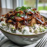A close-up view of a hearty pulled pork bowl featuring juicy shredded meat, fresh cilantro garnish, and tangy slaw on a bed of steaming grains.