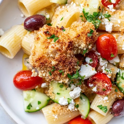 A close-up of Crispy Chicken Greek Pasta, showing golden fried chicken, al dente fusilli, and vibrant salad veggies in a zesty vinaigrette.