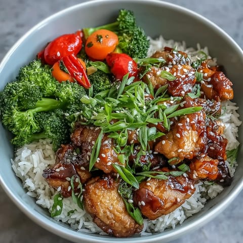 Bite-sized Honey Garlic Chicken glazed in sticky sauce is topped with green onions and sesame seeds beside colorful steamed vegetables and rice.
