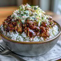 Golden-brown, tender pulled pork piled high on fluffy white rice, topped with crisp, creamy coleslaw and a drizzle of smoky BBQ sauce in a rustic ceramic bowl.